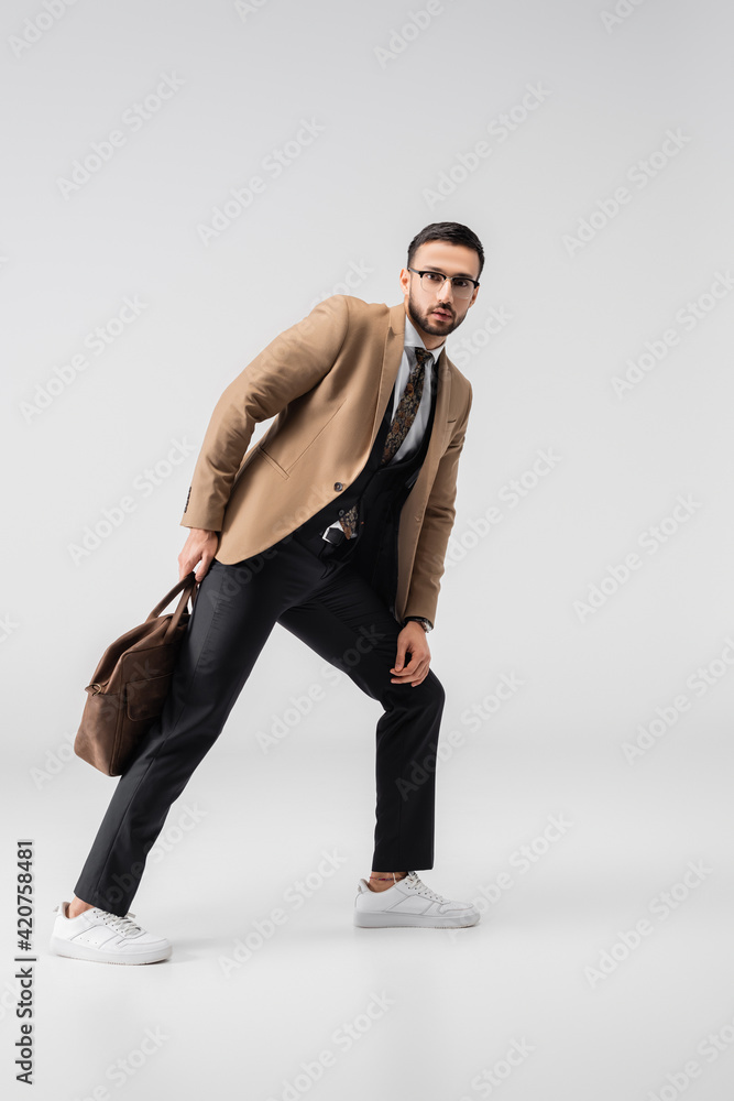 full length view of trendy muslim man looking at camera while posing with leather bag on grey full length view of trendy muslim man looking at camera while posing with leather bag on grey