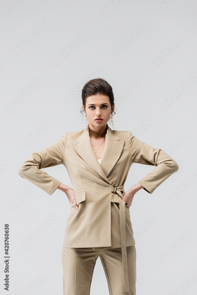 stylish woman in trendy pantsuit and pearl earrings posing with hands on hips isolated on grey