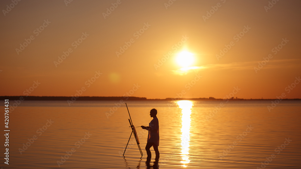 A young girl with a short haircut in lingerie draws a half-painting on a mulbert standing in the water at sunset of the lake. The artist paints a picture on the seashore. 