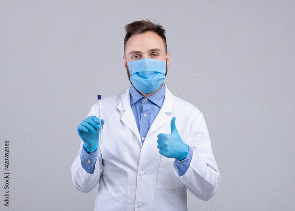 Portrait of doctor in mask and gloves holding covid-19 PCR test tube and showing thumb up gesture on grey background Portrait of doctor in mask and gloves holding covid-19 PCR test tube and showing thumb up gesture on grey background