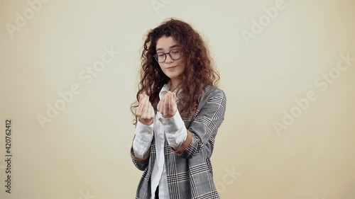 The beautiful employee shows with her hand the gesture of money, in order to receive a payment bonus. Cretaceous in gray jacket and white shirt, with glasses posing isolated on a beige background in