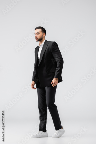 full length view of young arabian man in black, elegant suit and sneakers looking away on grey