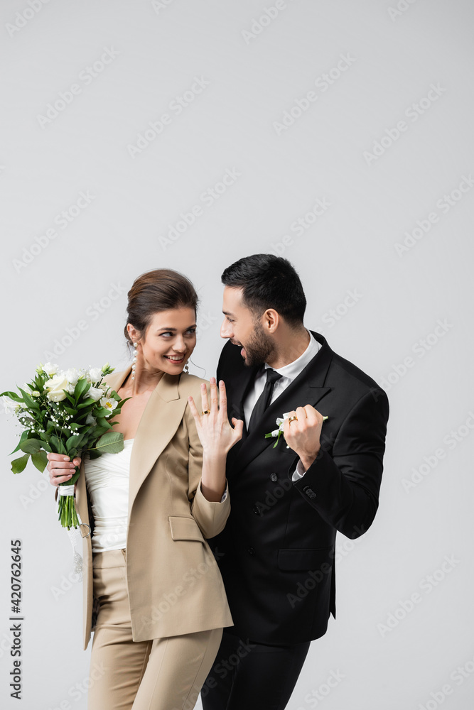 excited muslim groom showing win gesture while looking at cheerful bride isolated on grey
