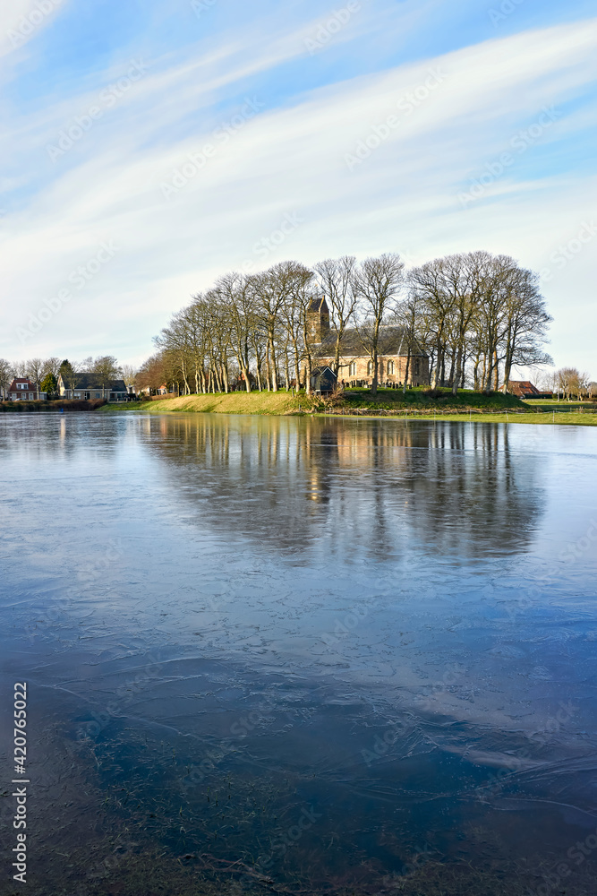 The 12th century romanesque Saint Nicholas Church on mound in Hijum Friesland with outdoor ice-skating rink in front during winter. Vertical image.