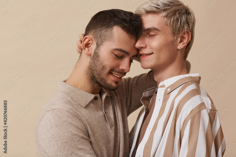 Minimal portrait of happy gay couple embracing lovingly while posing against beige background in studio Minimal portrait of happy gay couple embracing lovingly while posing against beige background in studio