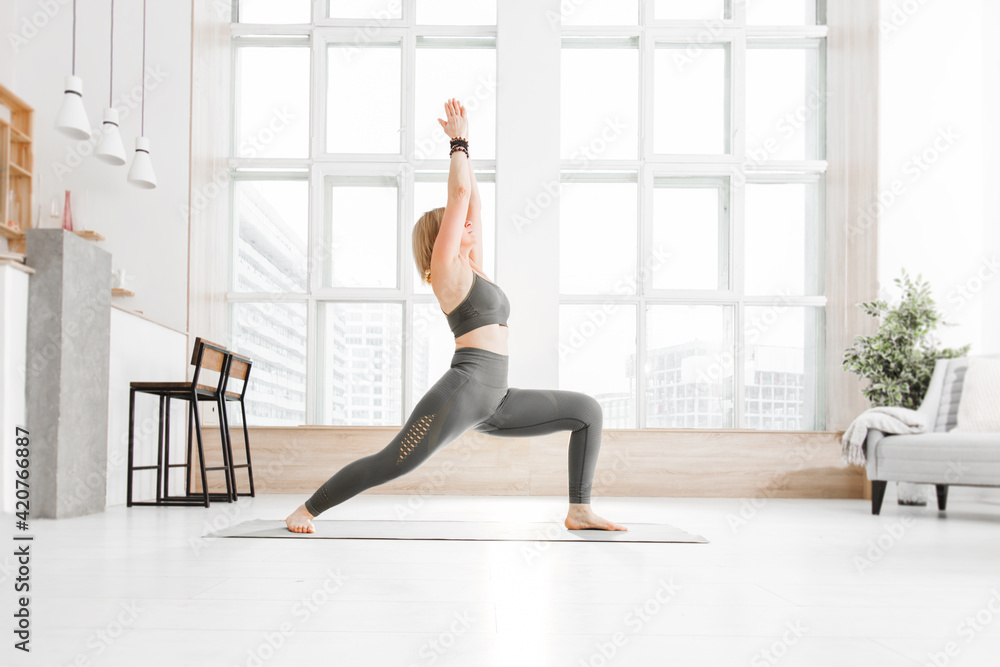 An adult woman practices Ashtanga Vinyasa yoga at home on the background of a window. Virabhadrasana pose An adult woman practices Ashtanga Vinyasa yoga at home on the background of a window. Virabhadrasana pose