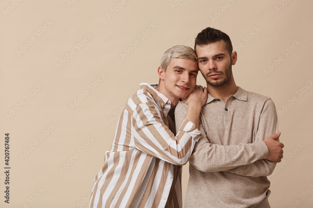 Minimal waist up portrait of happy gay couple looking at camera while posing against beige background in studio, copy space Minimal waist up portrait of happy gay couple looking at camera while posing against beige background in studio, copy space