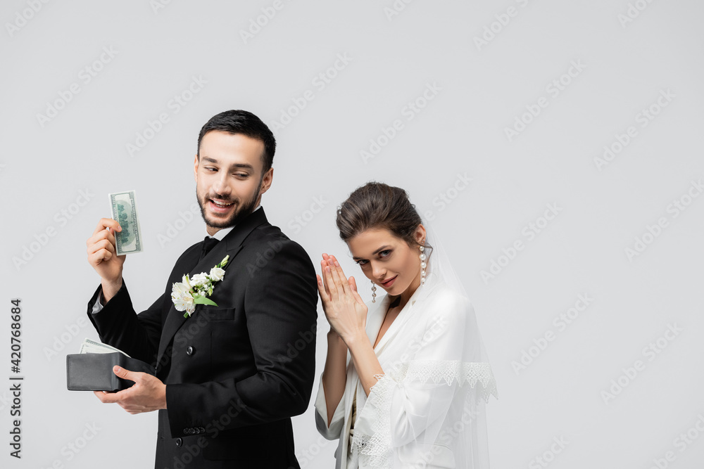 Muslim groom holding dollar banknote near tricky bride isolated on grey Muslim groom holding dollar banknote near tricky bride isolated on grey