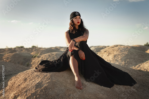 Woman in black dress and turban without shoes sits in desert