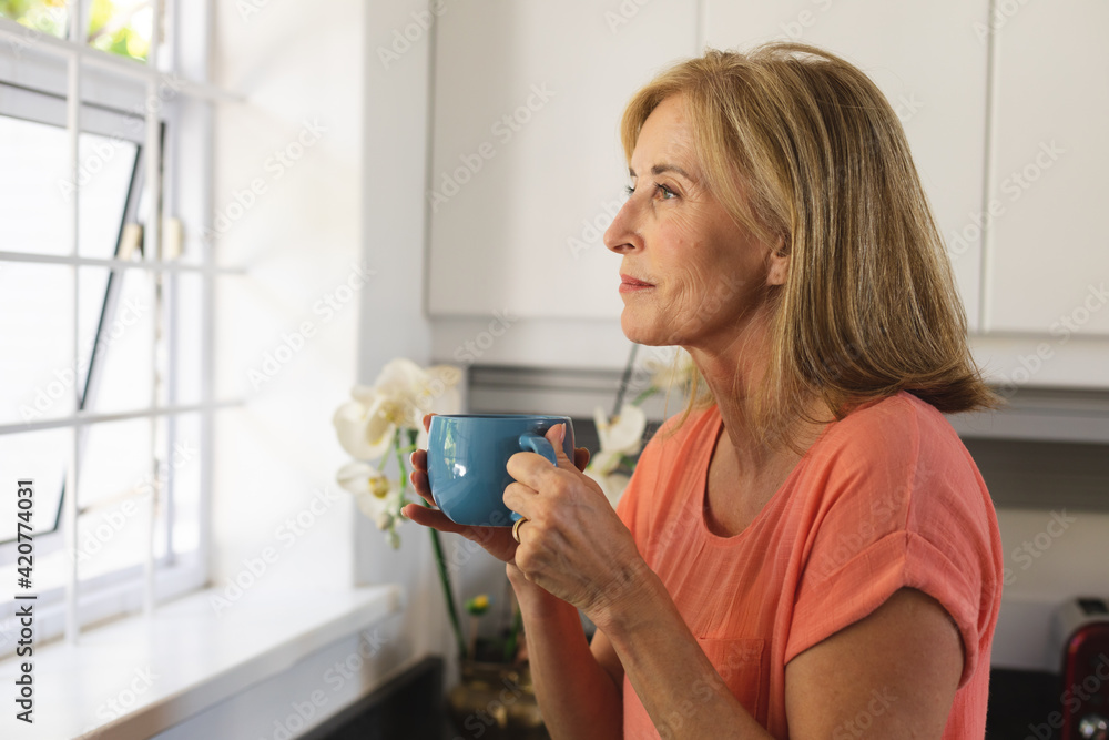 Caucasian senior woman standing by window drinking cup of coffee Caucasian senior woman standing by window drinking cup of coffee