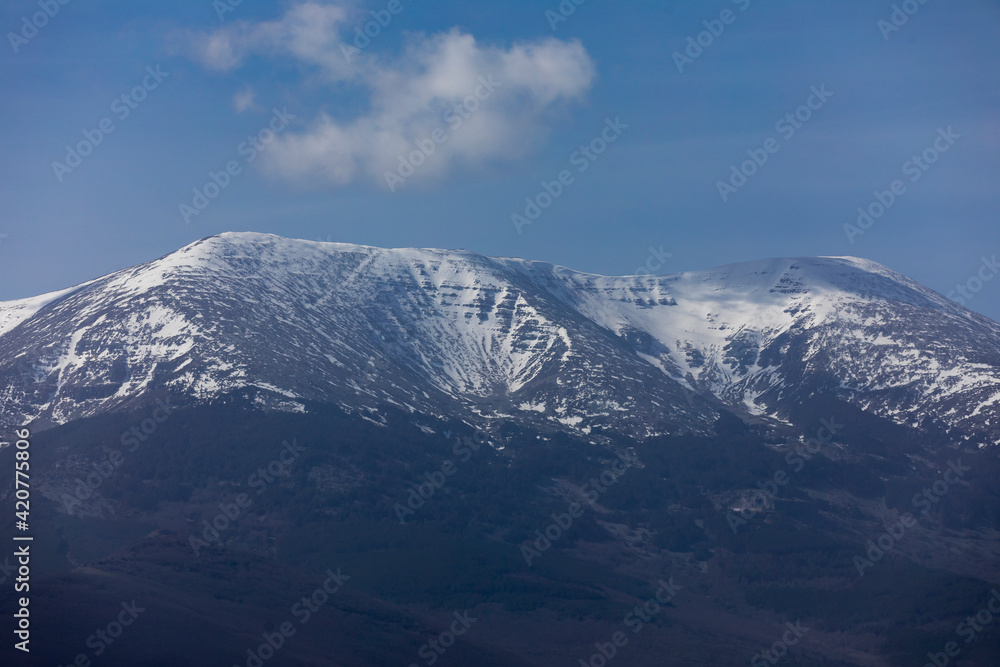 Photograph of Moncayo, the highest mountain in the Iberian System, covered with snow at its top, on a sunny winter day. On its slopes, dark forests of holm oaks and silent pines.