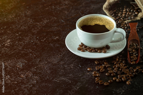 Hot black coffee for morning beverage menu in white ceramic cup with coffee beans roasted in burlap sack bag on dark grunge rustic table background. Flat lay with copy space.