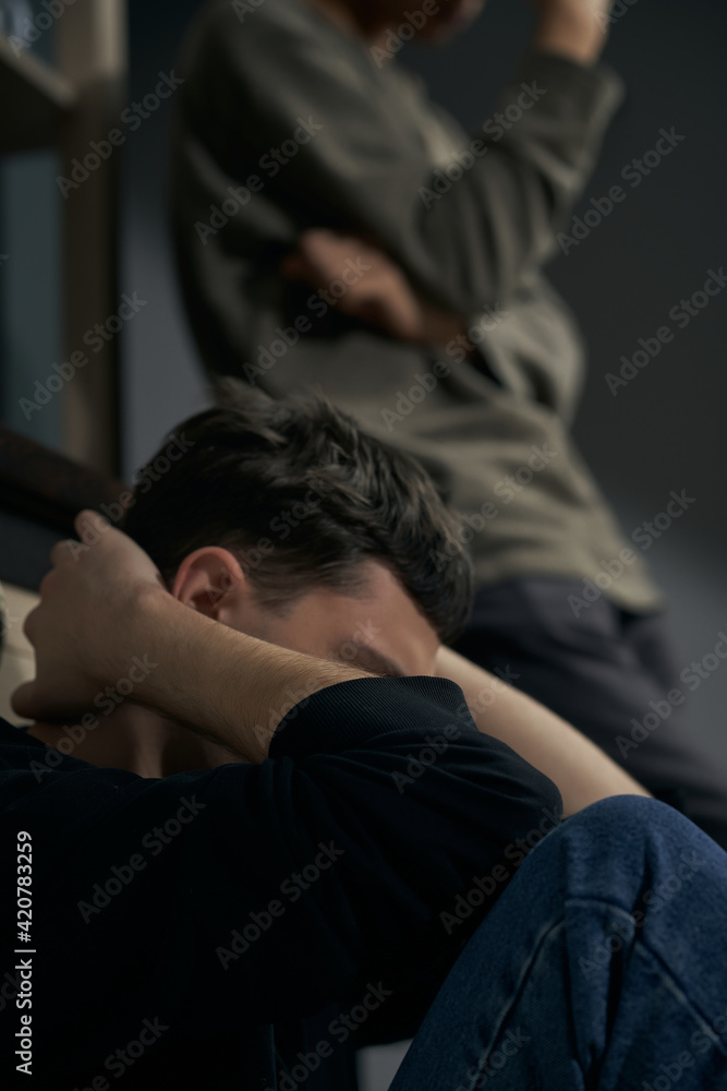 Staged photo illustrates problems and conflicts in gay couple relationships. Moment of showdown: young brooding and upset man is sitting in front of blurred figure of his partner. 