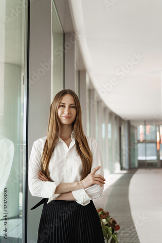 Vertical portrait of happy smiling businesswoman, young girl applied job in famous company, finished successful deal, cross hands chest confident pose, standing near window in company corridor