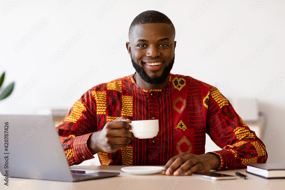 Happy Black Freelancer Guy In African Shirt Using Laptop And Drinking Coffee Happy Black Freelancer Guy In African Shirt Using Laptop And Drinking Coffee
