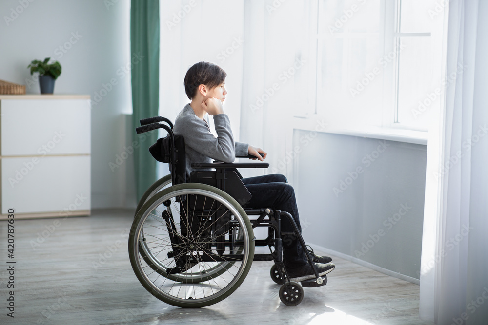 Side view of impaired teenage boy in wheelchair feeling desperate and lonely, looking out window at home Side view of impaired teenage boy in wheelchair feeling desperate and lonely, looking out window at home