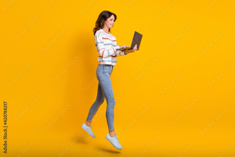 Full body profile side photo of young girl happy smile jump up use laptop chat type email isolated over yellow color background Full body profile side photo of young girl happy smile jump up use laptop chat type email isolated over yellow color background