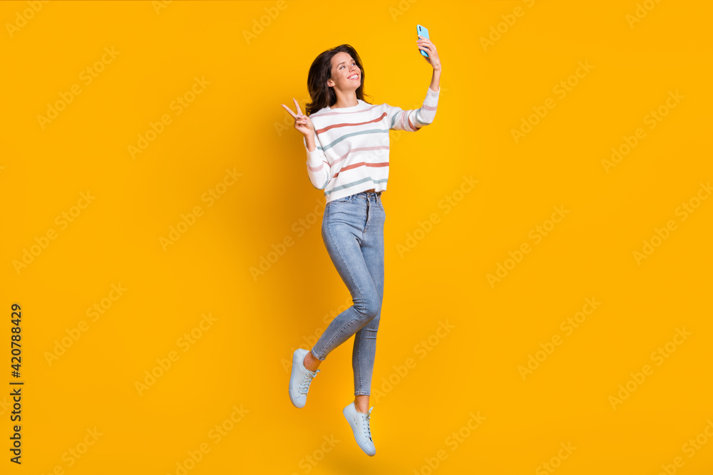 Full size photo of young girl happy smile jump up make take selfie show peace cool v-sign isolated over yellow color background Full size photo of young girl happy smile jump up make take selfie show peace cool v-sign isolated over yellow color background