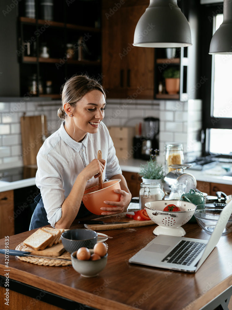 Young woman cooking in the kitchen. Beautiful woman following recipe on laptop and preparing delicious food.