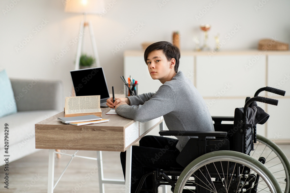 Disabled teen boy in wheelchair studying online with textbooks and laptop at home, mockup for design on pc screen Disabled teen boy in wheelchair studying online with textbooks and laptop at home, mockup for design on pc screen