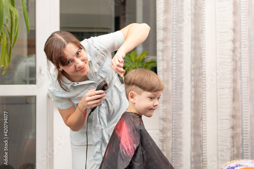 young mom hairdresser cuts her baby boy at home with hair clipper during quarantine. selective focus. portrait