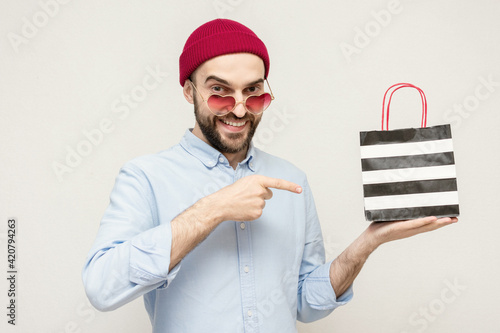 Bearded man points to the shopping bag, mman with the glasses in the shape of a heart shows buy, portrait, white background photo