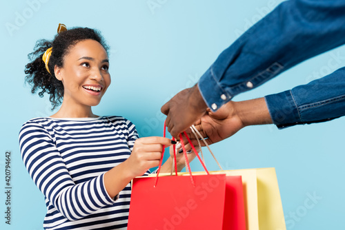 cheerful african american wife taking shopping bags from husband on blue