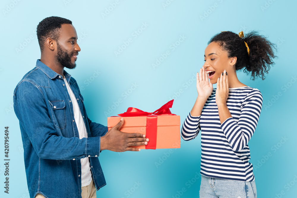 amazed african american woman near husband with big gift box on blue amazed african american woman near husband with big gift box on blue