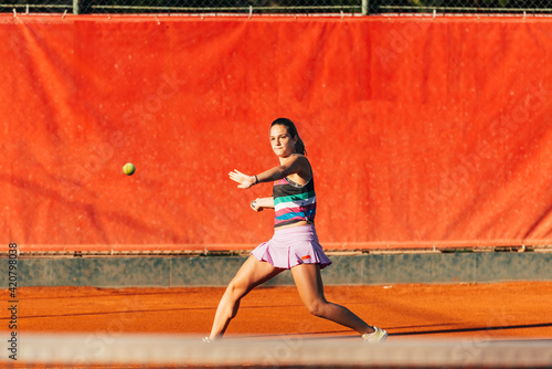 A young fit woman plays tennis outdoor on an orange tennis field early in the morning © qunica.com