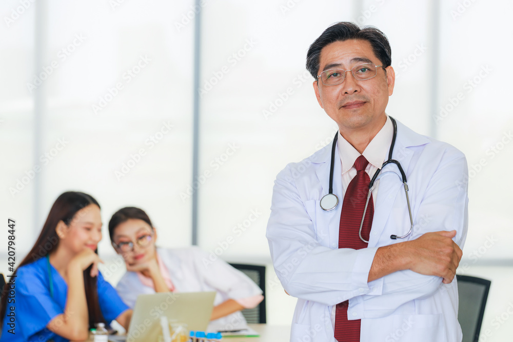 Portrait senior male doctor wearing eye glasses with background of young female doctor, and a young nurse with laptop are meeting in a meeting room in the hospital. Portrait senior male doctor wearing eye glasses with background of young female doctor, and a young nurse with laptop are meeting in a meeting room in the hospital.