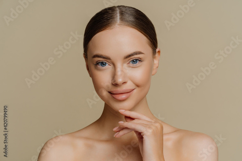 Headshot of beautiful brunette woman touches jawline looks directly at camera stands shirtless has natural beauty isolated over brown background. People wellness cosmetology skin care concept photo