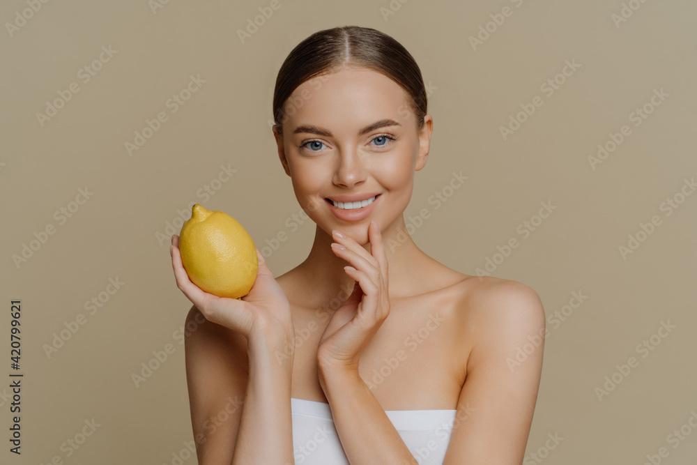 Cosmetology skin care and beauty concept. Pleased brunette young woman wrapped in bath towel holds lemon for making facial mask has refreshed clean skin after taking shower and spa procedures Cosmetology skin care and beauty concept. Pleased brunette young woman wrapped in bath towel holds lemon for making facial mask has refreshed clean skin after taking shower and spa procedures