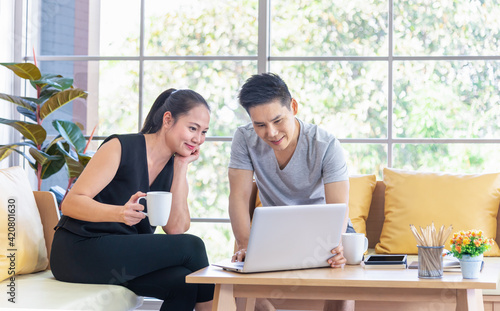 Beautiful young asian couple using laptop on the sofa at home. Young man working from home on laptop. Woman is smiling at her. Happy couple enjoying work from home