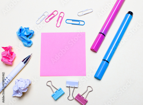 On a light background lies a pink sticker, a white pen, light blue and pink markers, paper clips. Top view with copy space. Flat lay