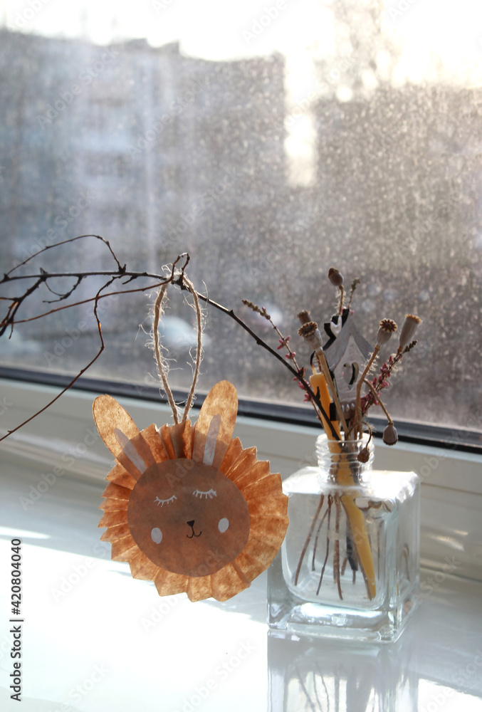 Easter bunny made of paper on a branch, composition on the window in the rays of the setting sun, dried flowers in a vase