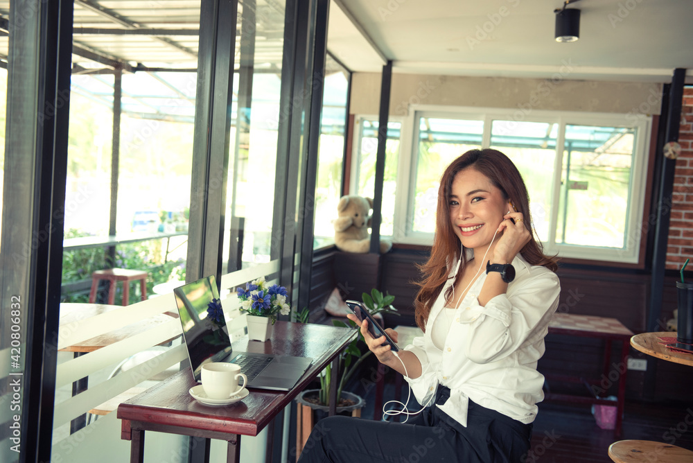 An Asian business woman sits in a coffee shop at a wooden table, drinks coffee and uses a smartphone on the table with a laptop. A woman holding a phone and looking at the screen An Asian business woman sits in a coffee shop at a wooden table, drinks coffee and uses a smartphone on the table with a laptop. A woman holding a phone and looking at the screen
