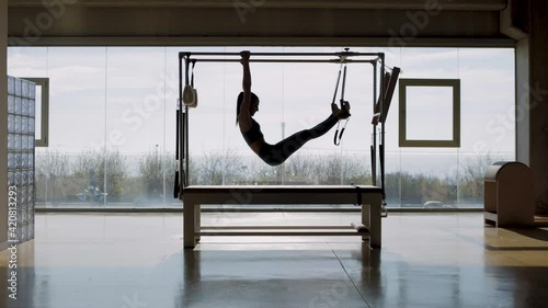 Tracking shot in slow motion of Fit female instructor hanging from the pilates machine, woman working out on the Pilates Trapeze Table in a big Fitness Studio with wide windows and street views.