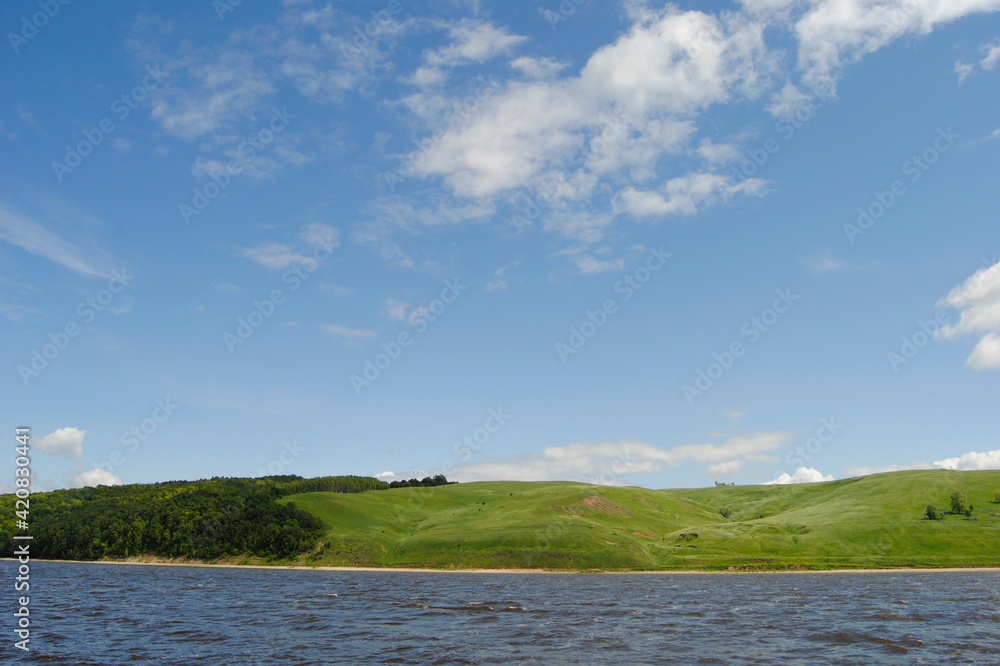 hilly green banks of a river or lake with forest on the background of a beautiful blue sky with clouds in summer