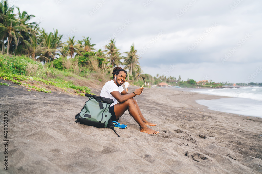 Happy ethnic man in earphones sitting on beach with smartphone