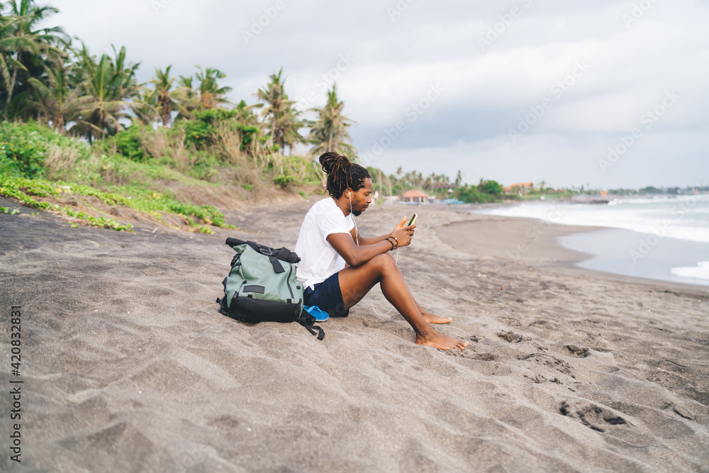 Ethnic man chilling on sandy beach and listening to music on smartphone