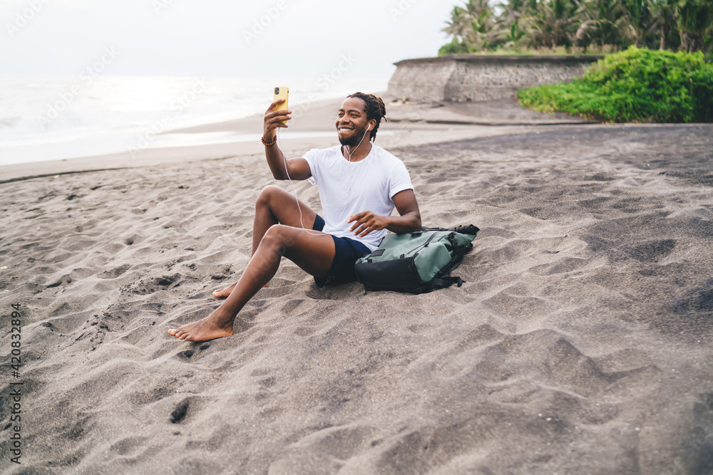 Joyful ethnic man sitting on sandy beach and making video call on smartphone