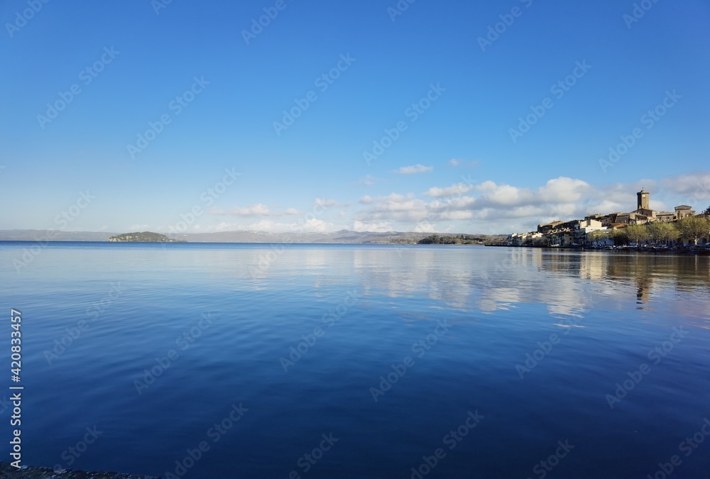 View of Lake Bolsena with the reflections of houses and clouds in the blue water rippled by the wind.