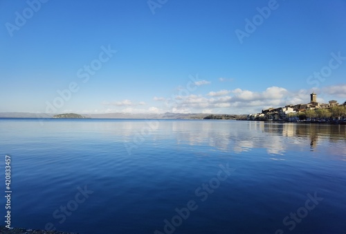 View of Lake Bolsena with the reflections of houses and clouds in the blue water rippled by the wind.