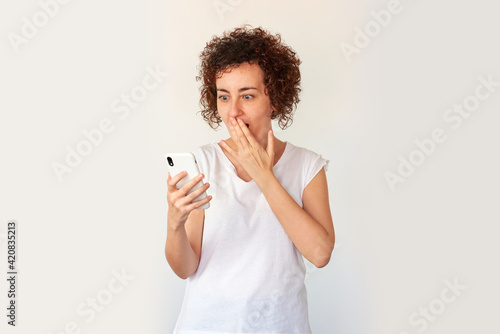 Curly haired caucasian young woman holding a smartphone with surprised face