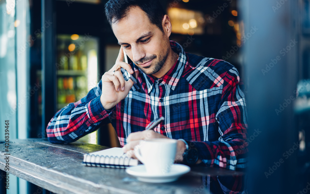 Caucaisan male writing notes during smartphone communication for discuss delivery details, handsome man in stylish shirt writing information in textbook while calling to partner for talking Caucaisan male writing notes during smartphone communication for discuss delivery details, handsome man in stylish shirt writing information in textbook while calling to partner for talking