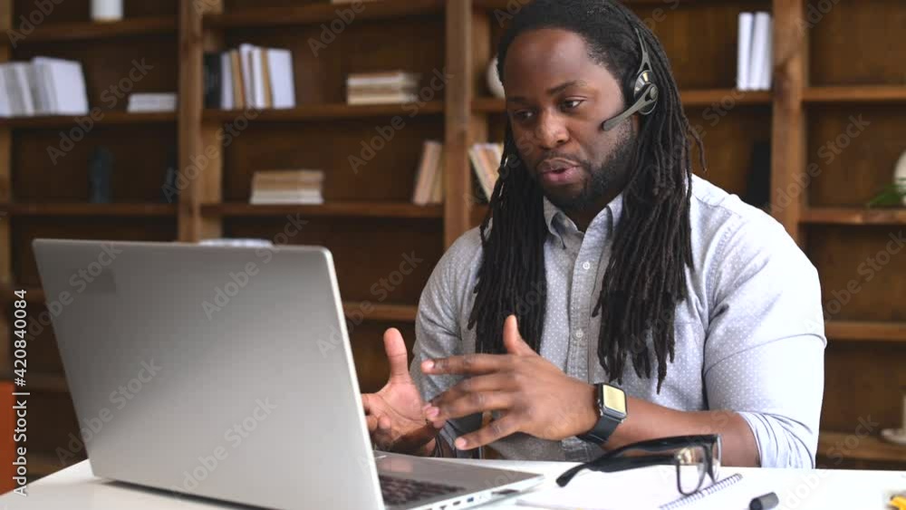 Diverse office employees concept. A focused African-American man with a dreadlocks wearing wireless headset is using laptop for online video conversation, talking with customers or coworkers online Diverse office employees concept. A focused African-American man with a dreadlocks wearing wireless headset is using laptop for online video conversation, talking with customers or coworkers online
