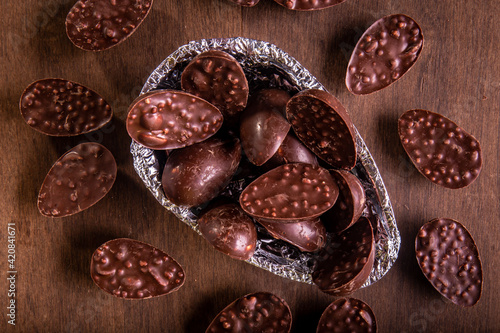 Top view of a close up of mini stuffed chocolate easter eggs inside of a half of an easter egg wrapped in an aluminium paper. Some mini stuffed easter eggs around it on a wooden table.