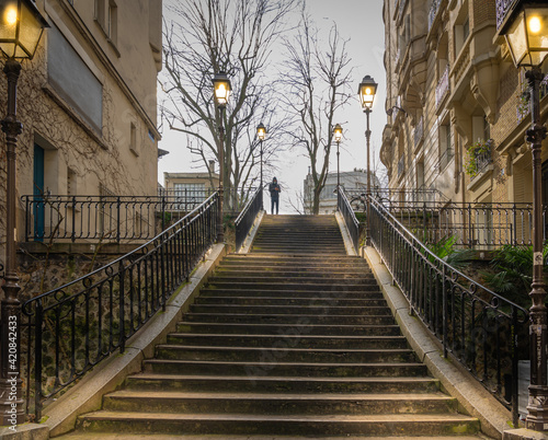 Fototapeta Naklejka Na Ścianę i Meble -  Paris, France - 02 26 2021: Montmartre district. Stone staircase with illuminated lanterns and a strange masked man.