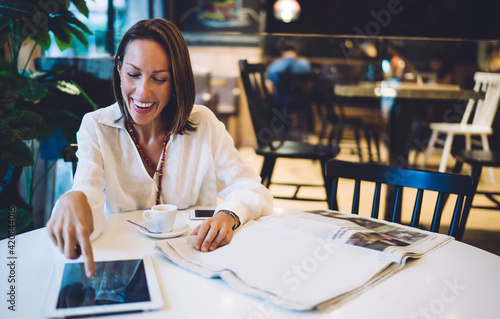 Cheerful female customer touching screen of gadget checking received email message and laughing, joyful blogger using digital tablet for online networking during morning time with fresh newspaper