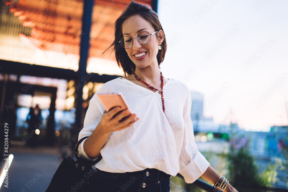 Happy Caucasian blogger connecting to 4g wireless for sharing travel publication via mobile application, cheerful woman in classic spectacles smiling while chatting via smartphone technology